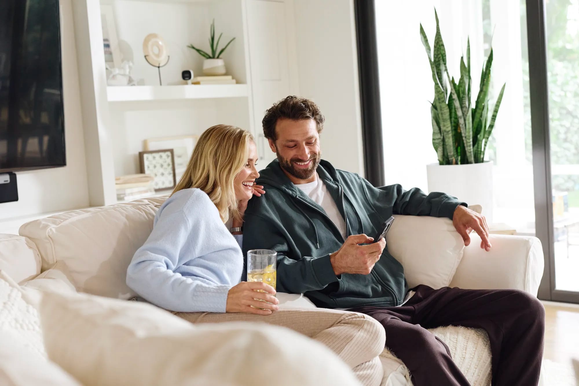A relaxed couple on a living-room sofa checking their home's environmental sensor alerts on a phone