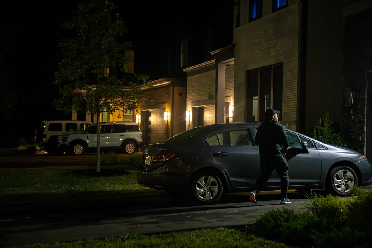 A person approaching a car at night outside a lit home, watched over by 24/7 monitoring