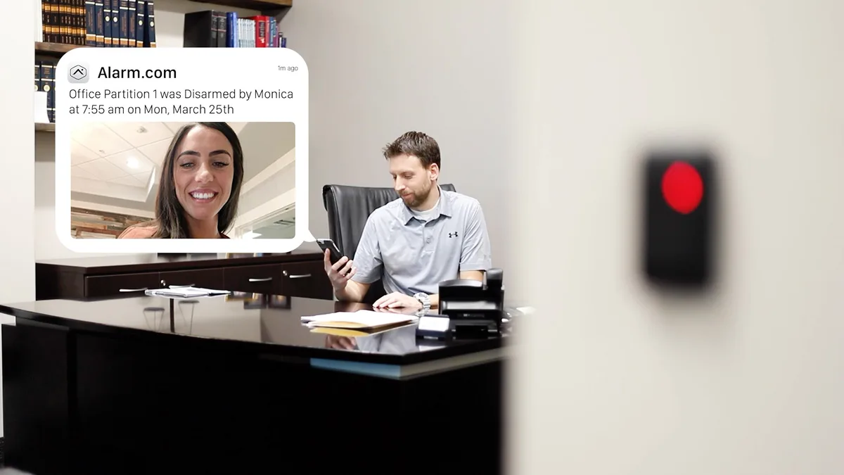 A business owner at his office desk receiving an Alarm.com mobile notification with video confirming an employee disarmed the office security system