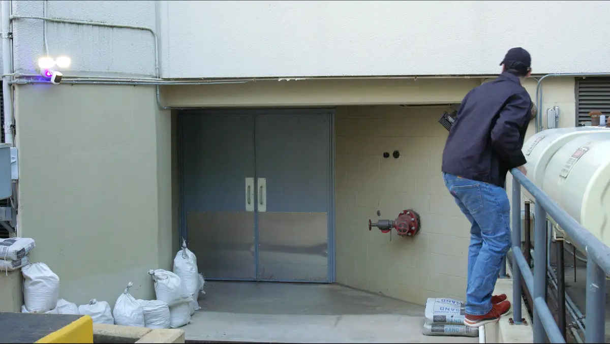 A man in a dark jacket and cap stepping up beside a commercial loading-dock door at dusk, watched by a wall-mounted security camera with active deterrent lights