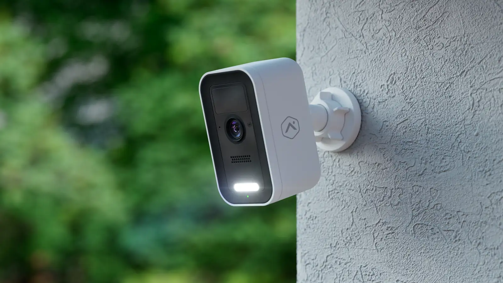 A white wall-mounted outdoor security camera covering a home's exterior, with lush greenery in the background