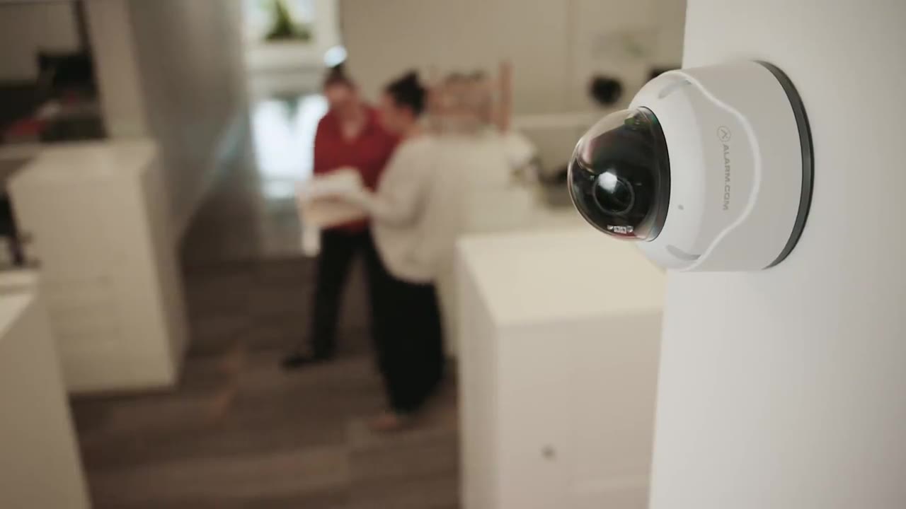 Alarm.com Prism dome security camera mounted on a wall watching over office workers in a bright modern workspace
