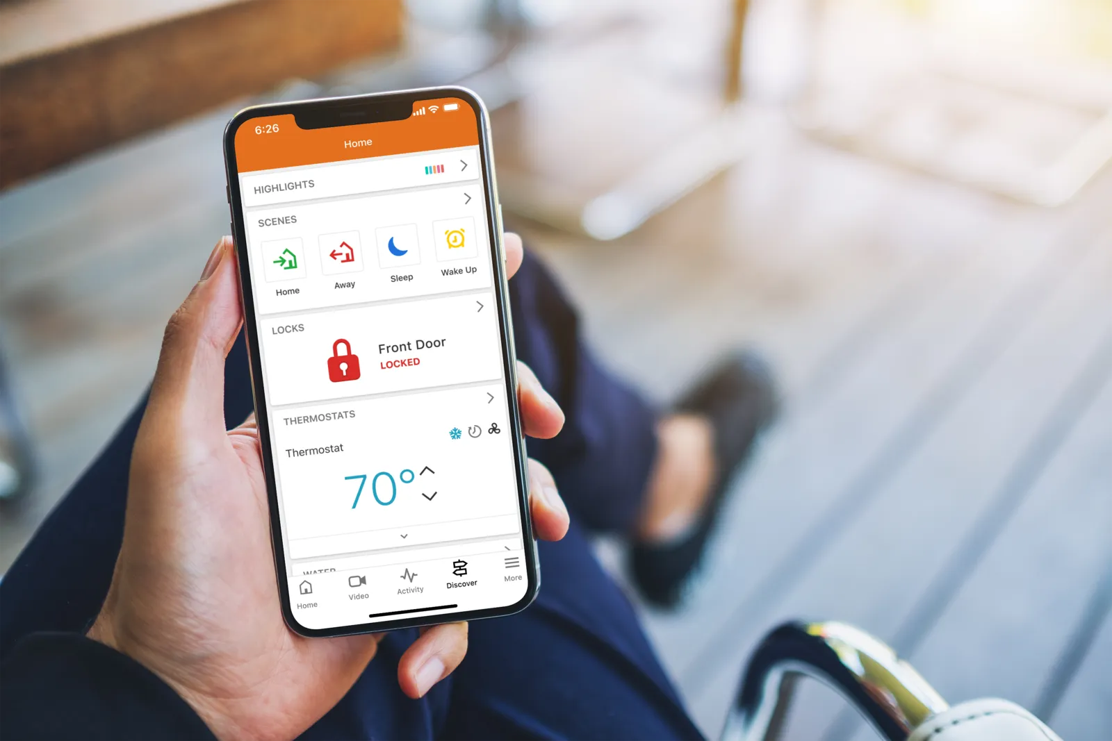 A hand holding a phone showing the Alarm.com app with scenes, front-door lock status, and thermostat controls for whole-home monitoring