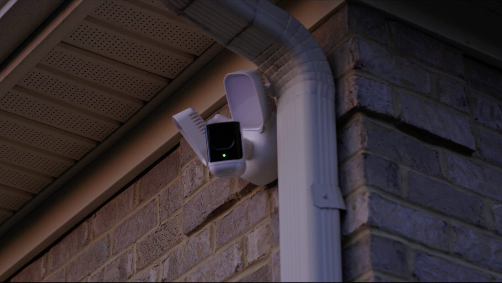An exterior floodlight security camera mounted under a home's roofline at dusk