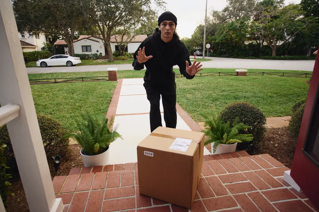 A hooded figure approaching a home's front door past a delivered package, watched over by 24/7 monitoring
