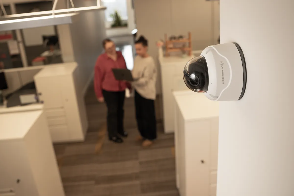 A wall-mounted Alarm.com dome security camera monitoring an open office while two colleagues talk in the background